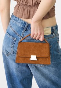 Brown suede handbag with a structured design, featuring a silver clasp and a short handle, held near high-waisted blue denim jeans.