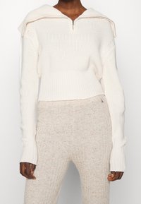 Weißer, gerippter Strickpullover mit einem Reißverschlusskragen und verlängerten Ärmeln, kombiniert mit beigen, strukturierten, gerippten Hosen.