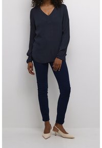Blouse manches longues bleu marine avec un col en V et une patte de boutonnage à l'avant, associée à un jean skinny bleu foncé et des escarpins à bride arrière beige clair.