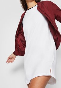 Röd bomberjacka med ribbade muddar, bärs öppen över en vit t-shirtklänning med sidofickor. Släta material, modern avslappnad stil.