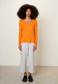 Oranje shirt met lange mouwen en een ronde hals, gecombineerd met grijze cropped joggingbroek en zwarte instapschoenen, op een getextureerde vloer.