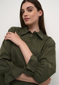 Olive green blouse with a button-up front, structured collar, and puffed sleeves with gathered cuff detail. Smooth, lightweight fabric.