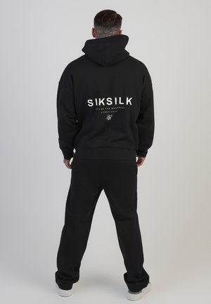 Mand set bagfra iført en sort hættetrøje og bukser med hvidt logo "SIKSILK" på ryggen, stående mod en ensfarvet lys baggrund.