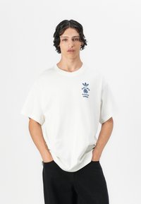 White cotton t-shirt with short sleeves, featuring a blue graphic print of a logo and design details on the left chest. Black pants.