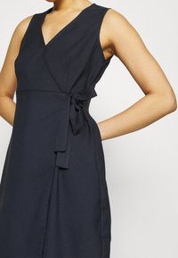 Bezramenné tmavě námořnické šaty na zavazování s véčkovým výstřihem a detaily na zavázání v pase, nošené osobou s jednou rukou na boku na jednobarevném pozadí.