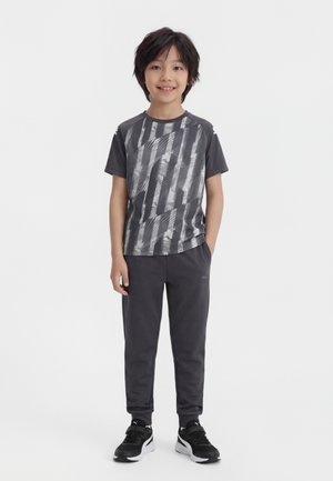 Αγόρι που στέκεται και χαμογελά φορώντας γκρι γραφικό μπλουζάκι, σκούρο γκρι παντελόνι και μαύρα και λευκά αθλητικά παπούτσια σε λευκό φόντο.