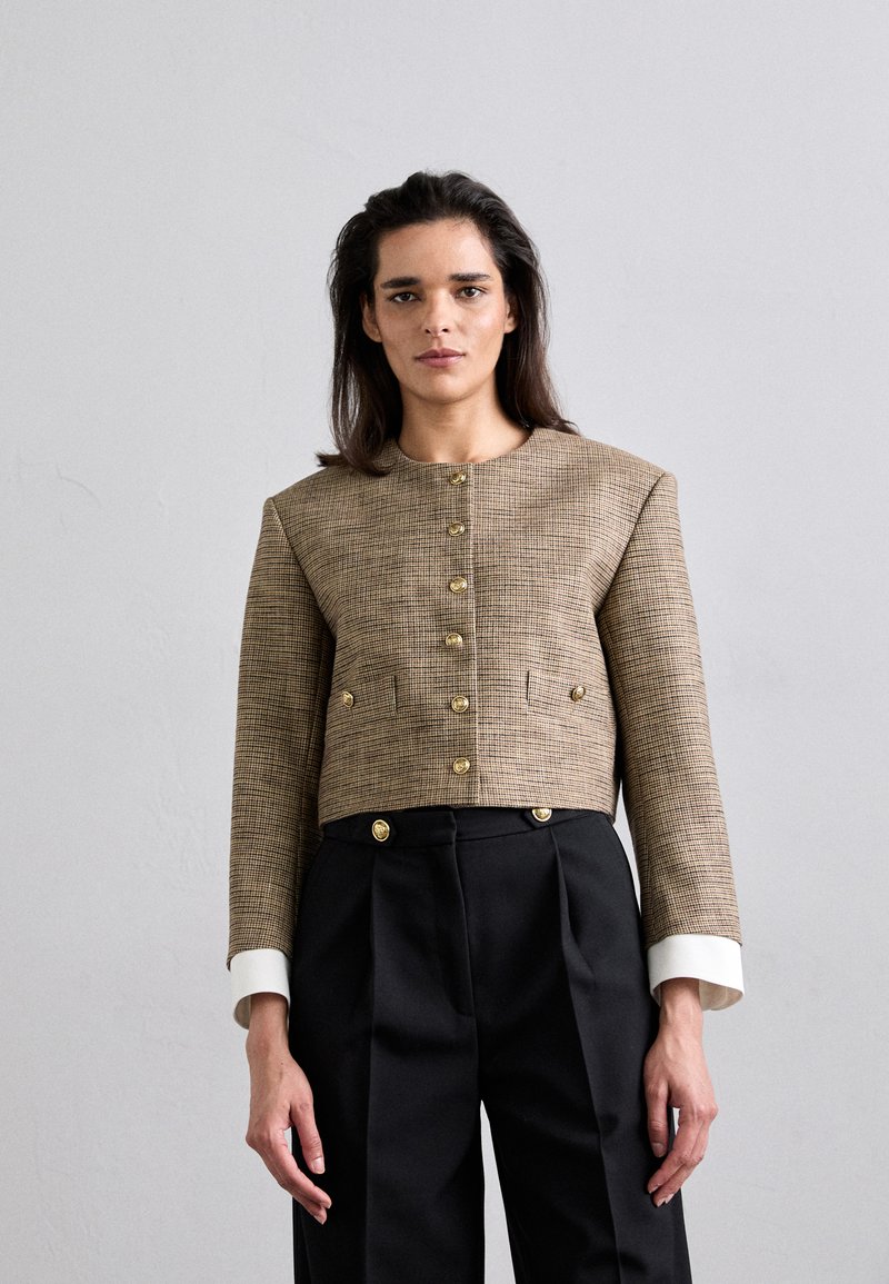 Geknipte, geweven blazer in een bruin ruitpatroon met gouden knopen, voorzien van een ronde halslijn en lange mouwen met witte manchetten. Gecombineerd met zwarte broeken.
