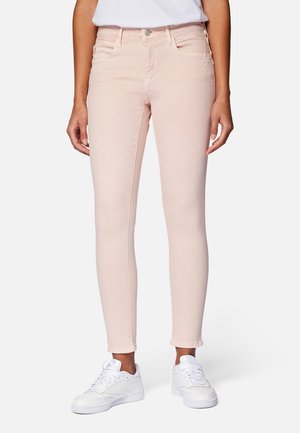 Personne portant un jean skinny rose clair et des baskets blanches, debout devant un fond clair et uni.