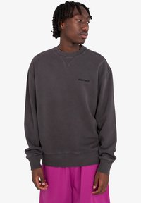 Grauer Rundhalsausschnitt-Sweatshirt in entspannter Passform, mit geripptem Kragen und Bündchen, und einem kleinen schwarzen Logo auf der Vorderseite. Getragen mit lila Shorts.