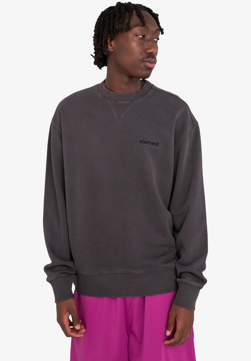 Felpa grigia a girocollo in un fit rilassato, con collo e polsini a coste, e un piccolo logo nero sulla parte anteriore. Indossata con pantaloni corti viola.