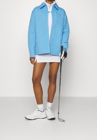 Světle modrá prošívaná bunda s límcem, bílé golfové šaty, bílé tenisky a golfová hůl. Obsahuje texturované vzory a kontrastní akcenty.