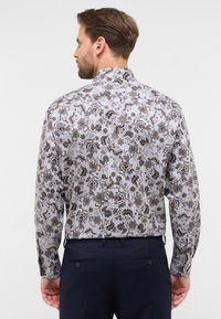 Bluse mit floralem Muster in Hellgrau mit dunklen Braun- und Beigemotiven. Verfügt über einen klassischen Kragen und lange Ärmel, kombiniert mit dunklen Hosen.