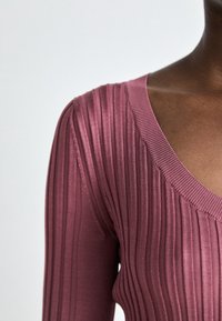 Rippenshirt mit tiefem V-Ausschnitt in Mauve und langen Ärmeln. Der Stoff hat eine weiche Textur und eine figurbetonte Silhouette, die vertikale Linien betont.