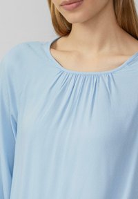 Hellbluse mit rundem Ausschnitt und gerafften Details an der Brust. Weicher Stoff mit glatter Textur und langen Ärmeln.