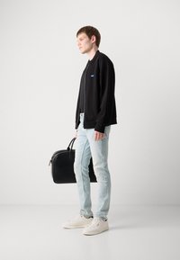 Juodas zip-up megztinis, šviesiai mėlynos pablukusios džinsinės kelnės ir baltos sportinės bateliai. Laiko juodą odinį krepšį, stovėdamas prieš neutralų foną.
