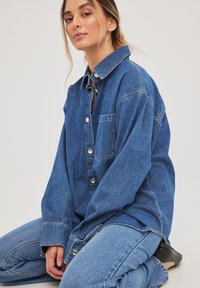 Camisa de denim azul medio con botones en el frente, collar tapered y un bolsillo en el pecho. Usada con jeans azules a juego.