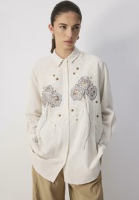 Light beige button-up shirt made of linen, featuring floral embroidery in gray and brown with textured stitching details. Loose fit, long sleeves.