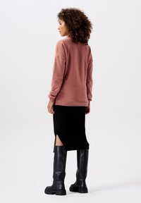 Rosa Sweatshirt mit lässiger Passform, kombiniert mit einem schwarzen, gerippten Rock mit seitlichen Schlitzen. Das Model trägt kniehohe schwarze Lederstiefel.