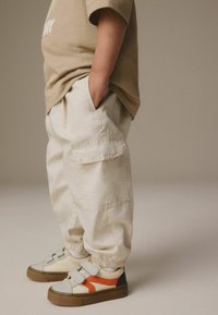 Beige cargo bukser med sidelommer, elastiske manchetter og en løs pasform, parret med beige og orange sneakers med velcro-stropper.