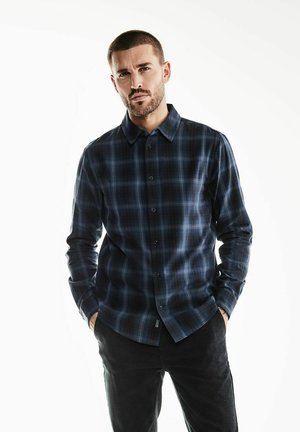 Geruite lange mouwen blouse in donkerblauw en zwart, gemaakt van zacht materiaal. Voorzien van een knopsluiting op de kraag en een knoopsluiting aan de voorkant.