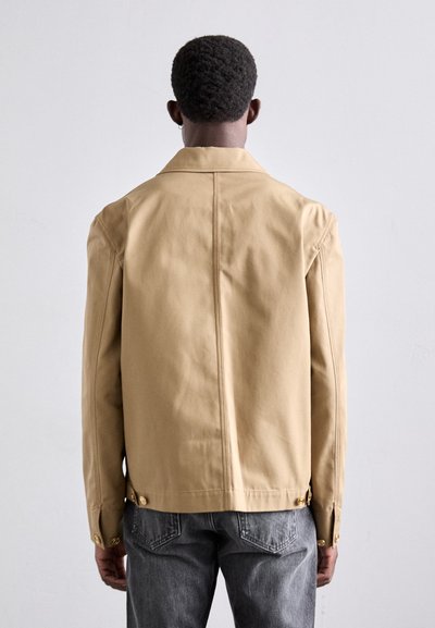 Beige jacket with a collared neck, featuring visible stitching and gold buttons at the cuffs. Worn over gray jeans, viewed from the back.