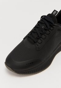 Baskets de sport noires avec une tige texturée, lacets fins noirs, semelle en caoutchouc et marquage en relief sur le côté et sur la languette.