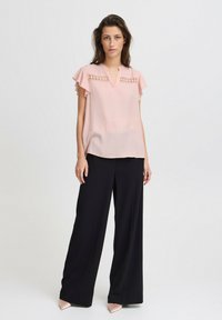Rosa Bluse mit kurzen Rüschensäumen, V-Ausschnitt und aufwändigen Spitzenverzierungen an der Schulter. Kombiniert mit schwarzen weit geschnittenen Hosen.