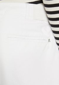 Białe spodnie z tylną kieszenią na zamek, wykonane z gładkiej tkaniny, z widoczną metką marki i kontrastującym paskiem w paski w talii.