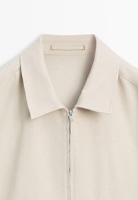 Beige Hemd mit Kragen und sichtbarem silbernen Reißverschluss an der vorderen Mitte, aus strukturiertem Stoff gefertigt.