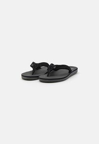 Des tongs noires avec une sangle texturée comportant un nœud décoratif, une semelle plate avec un minimum de profil et un design simple et fonctionnel.