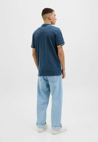 Navyblaues Polohemd mit kurzen Ärmeln und cremefarbenen Akzenten, kombiniert mit lässigen hellblauen Jeans. Vor einem schlichten weißen Hintergrund.