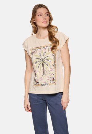 Mujer vistiendo una camiseta de manga corta beige con un detallado estampado de palmeras y flores, combinada con pantalones sueltos de color azul oscuro.