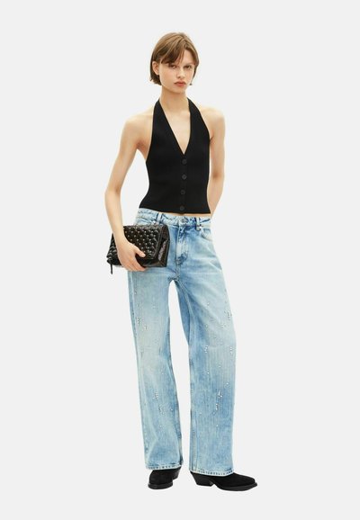 Vrouw in een zwarte haltertop met knopen, lichtblauwe, versierde wijd gesneden jeans, zwarte enkellaarsjes, met een zwart gestikte clutch. 
