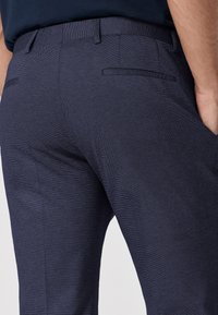 Pantaloni sartoriali blu navy in tessuto texture, con due tasche posteriori e una cintura liscia. Dettaglio a righe orizzontali sottili visibile.