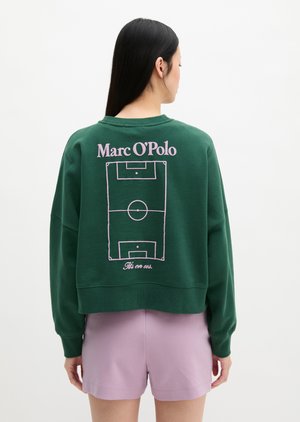 Žena s dugom tamnom kosom stoji okrenuta leđima, nosi zelenu majicu dugih rukava s ružičastim dizajnom nogometnog terena i tekstom "Marc O'Polo" na leđima.