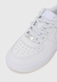 Baskets blanches avec une tige en cuir lisse, fermeture à lacets, boîte à orteils perforée et un accent logo métallique sur le côté. Semelle texturée.