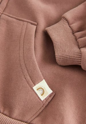 Tissu de sweat-shirt beige clair avec une poche et des poignets côtelés. Une petite étiquette avec un logo est cousue sur la poche. Texture douce.