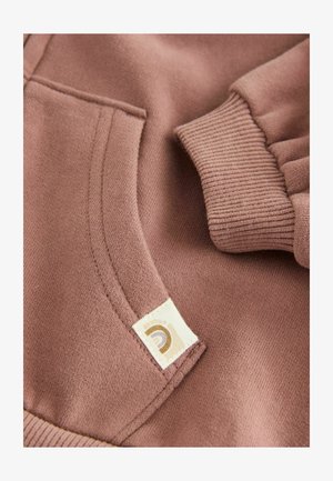 Tessuto di felpa beige chiaro con tasca e polsini a coste. Un piccolo etichetta con un logo è ricamata sulla tasca. Texture morbida.