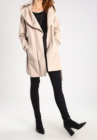 Beige trench coat with a large hood, button closure, and side pockets, worn over a black top and black skinny pants with black heels.