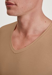 Beige v-hals t-shirt met korte mouwen, zachte textuur en subtiele stikdetails rond de halslijn, getoond in een close-up van een nek en borst.