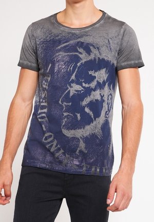 Mann trägt ein grau-navy T-Shirt mit einem großen abstrakten skizzenhaften Profil eines Männergesichts und teilweisem Text auf der Vorderseite.