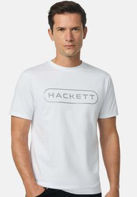 Camiseta de algodón blanca con un gran logo gris "HACKETT" en la parte delantera. Mangas cortas y cuello redondo. Textura suave.