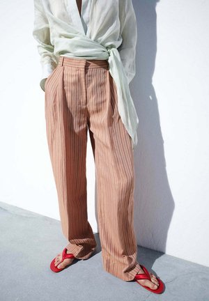 Pantalones anchos de color rosa claro con rayas finas combinados con una blusa cruzada blanca transparente y sandalias rojas de dedos sobre suelo de hormigón.