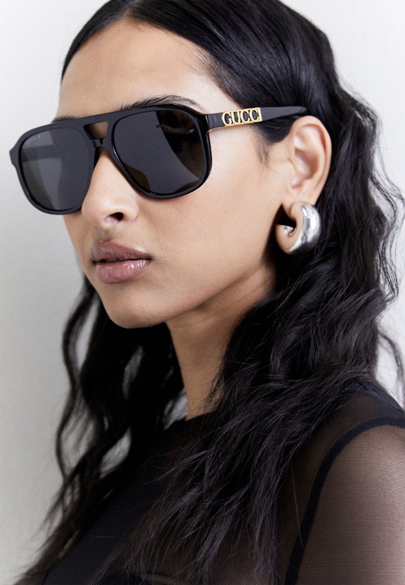 Lunettes de soleil rectangulaires noires avec des verres teintés et le logo "GUCCI" doré sur le côté ; design de monture élégant, portées avec un haut noir transparent.