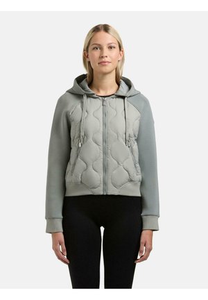 Kvinde med blondt hår iført en lysegrå quiltet hættetrøje med lynlås og snore samt sorte leggings, stående foran en ensfarvet hvid baggrund.