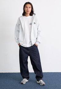 Felpa grigia con zip e coulisse regolabili, indossata sopra una t-shirt bianca a girocollo. Jeans blu scuro a vestibilità rilassata e sneakers grigie completano il look.