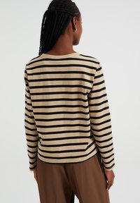 Žena s pletenými vlasy, oblečená v béžovém a černém pruhovaném tričku s dlouhým rukávem a hnědých kalhotách, otočená zády k jednobarevnému pozadí.