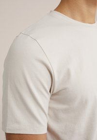 Tricou cu mânecă scurtă din țesătură bej deschis. Guler rotund cu textură netedă, cusătură vizibilă pe umăr și tiv bine finisat.