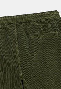 Pantalon en velours côtelé vert olive montrant la ceinture arrière, la couture latérale, et une poche passepoilée unique avec un petit logo circulaire au-dessus.