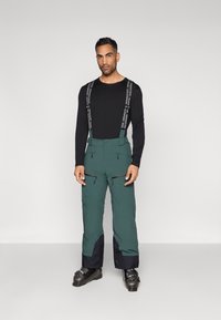 Calças de esqui verdes com suspensórios pretos ajust�áveis, bolsos com fecho éclair e uma parte inferior em tom azul marinho contrastante, combinadas com botas de esqui pretas.